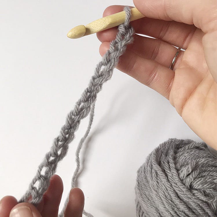 A person is holding a crochet hook to a grey yarn, working on a pattern.