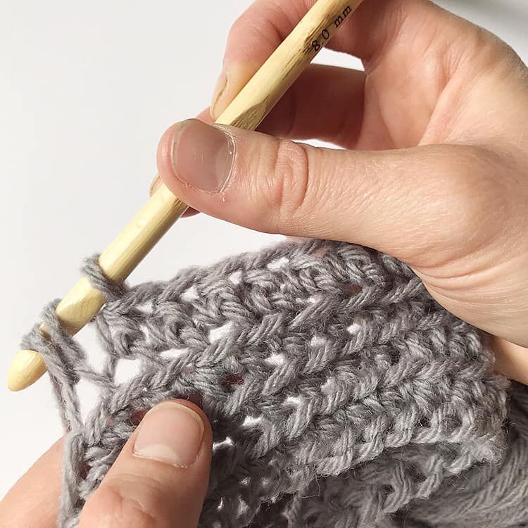 A person is using a crochet hook to knit a grey sweater pattern.