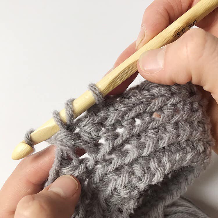 A person is using a wooden crochet hook to knit a grey sweater with a unique pattern.