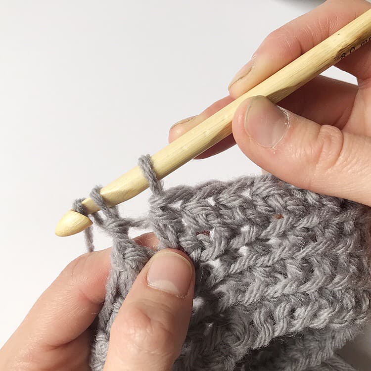 A person using a crochet hook to knit a grey sweater with a pattern.