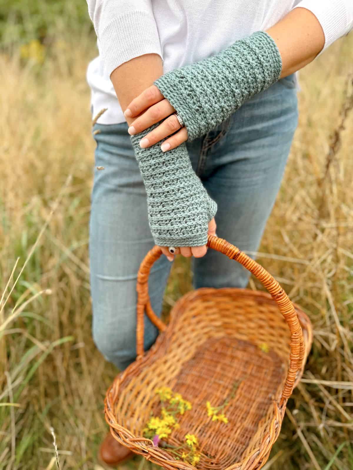 Person wearing fingerless gloves with a delicate crochet pattern, holding an empty wicker basket with a few yellow flowers, standing outside in a grassy area.