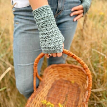 Person wearing light blue crocheted fingerless gloves, holding an empty wicker basket in a field, with green plants visible in the background. The intricate fingerless gloves crochet pattern complements the natural scenery beautifully.