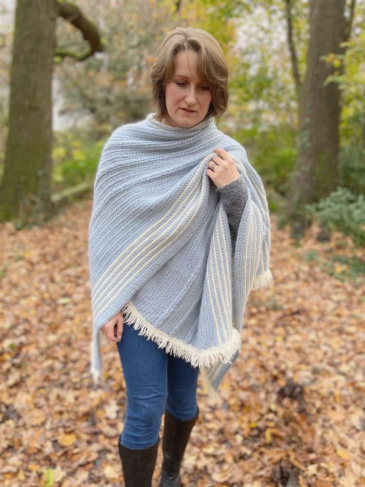 Woman in woods wearing jeans, boots and crochet wrap like a blanket.