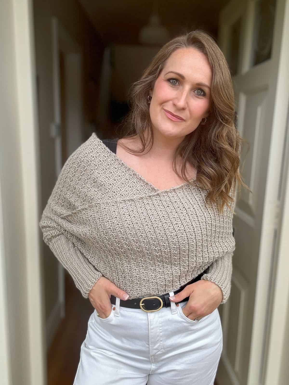 A woman with wavy brown hair stands indoors, wearing a beige off-shoulder crochet sweater scarf and white pants, her hand in her pocket while showcasing a scarf with sleeves crochet pattern draped elegantly over her shoulder.