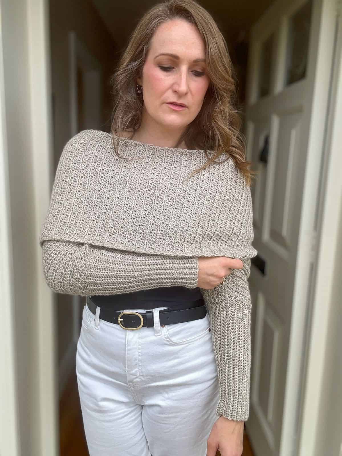 A woman with wavy hair, wearing a beige crochet sweater scarf and white pants, stands in a doorway with a pensive expression, contemplating a new scarf with sleeves crochet pattern.