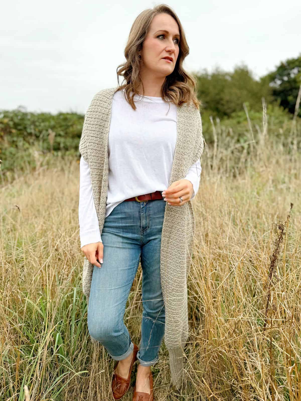 A woman stands in a grassy field wearing a white long-sleeve shirt, blue jeans, a long crochet scarf, and brown shoes. She has one hand in her pocket and is looking to the side, showcasing her unique scarf with sleeves crochet pattern that adds a touch of individuality to her outfit.