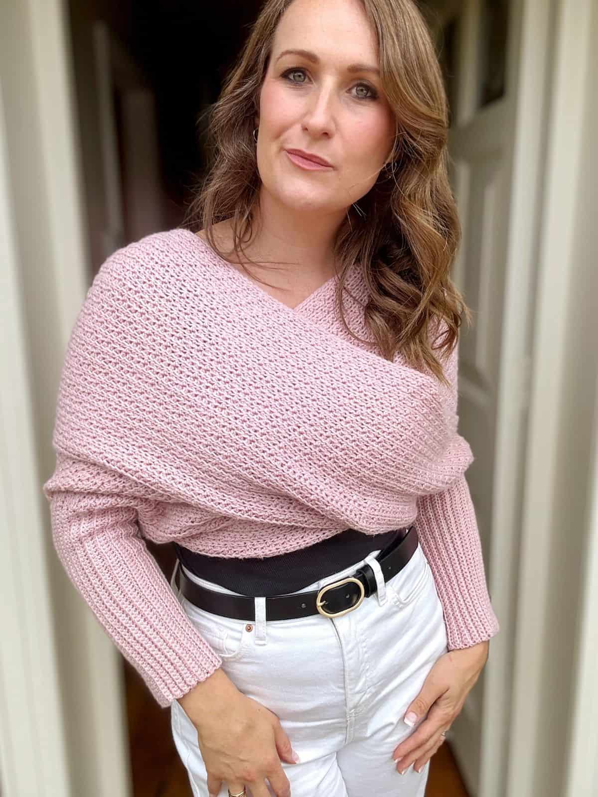 A woman stands indoors wearing a pink crochet sweater scarf with white pants and a black belt, looking directly at the camera, her outfit complemented by a scarf with sleeves crochet pattern.