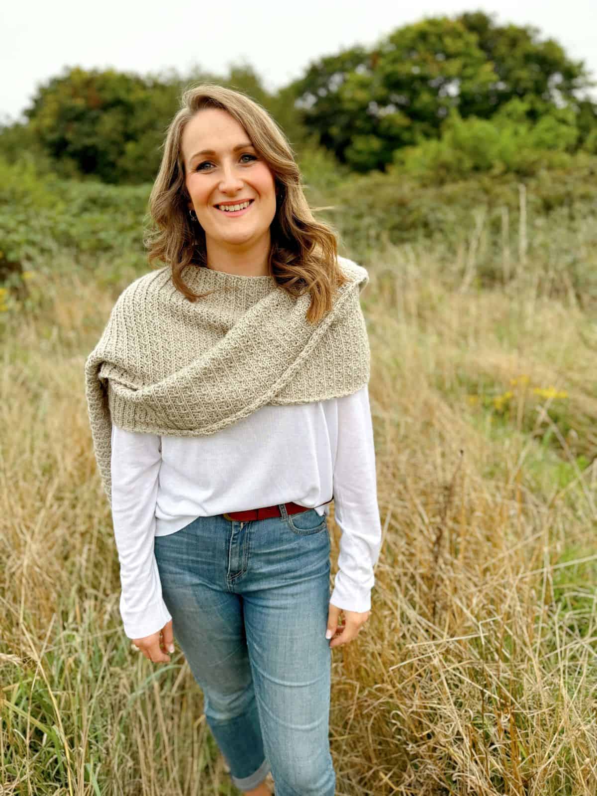 Woman in a white long-sleeve shirt, blue jeans, and a beige crochet shawl in grassy field with trees in the background, smiling at the camera. Her shawl appears to follow a beautiful crochet pattern, adding an elegant touch to her serene setting.