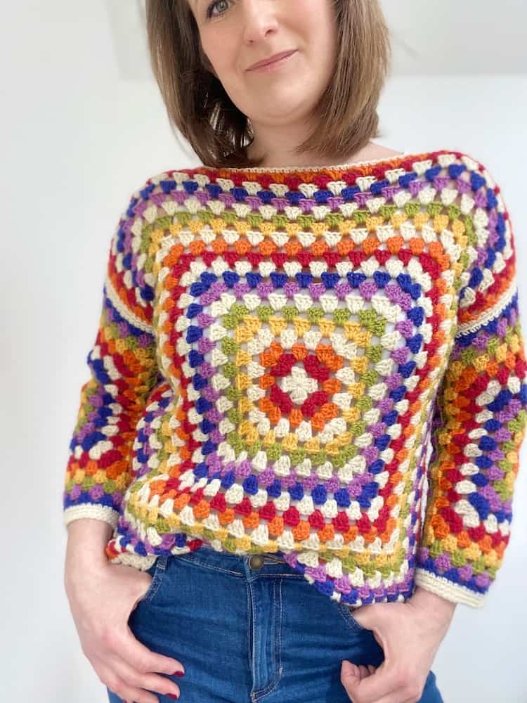 Woman wearing rainbow granny square sweater with hands in jeans pockets.