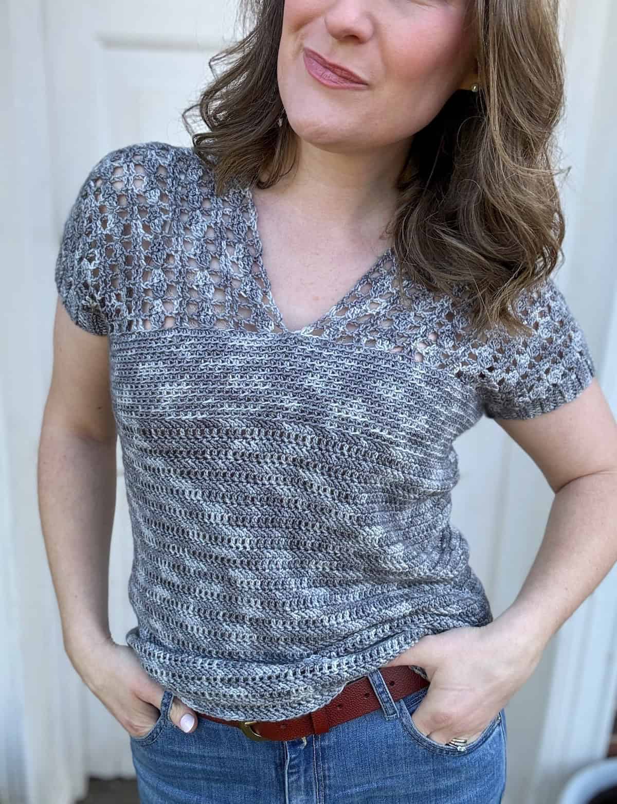 A person standing with hands in pockets, wearing a gray v neck crochet top and blue jeans, against a white background.