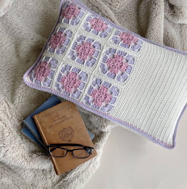 floral granny square cushion in pink, lilac and white next to a pile of books and glasses