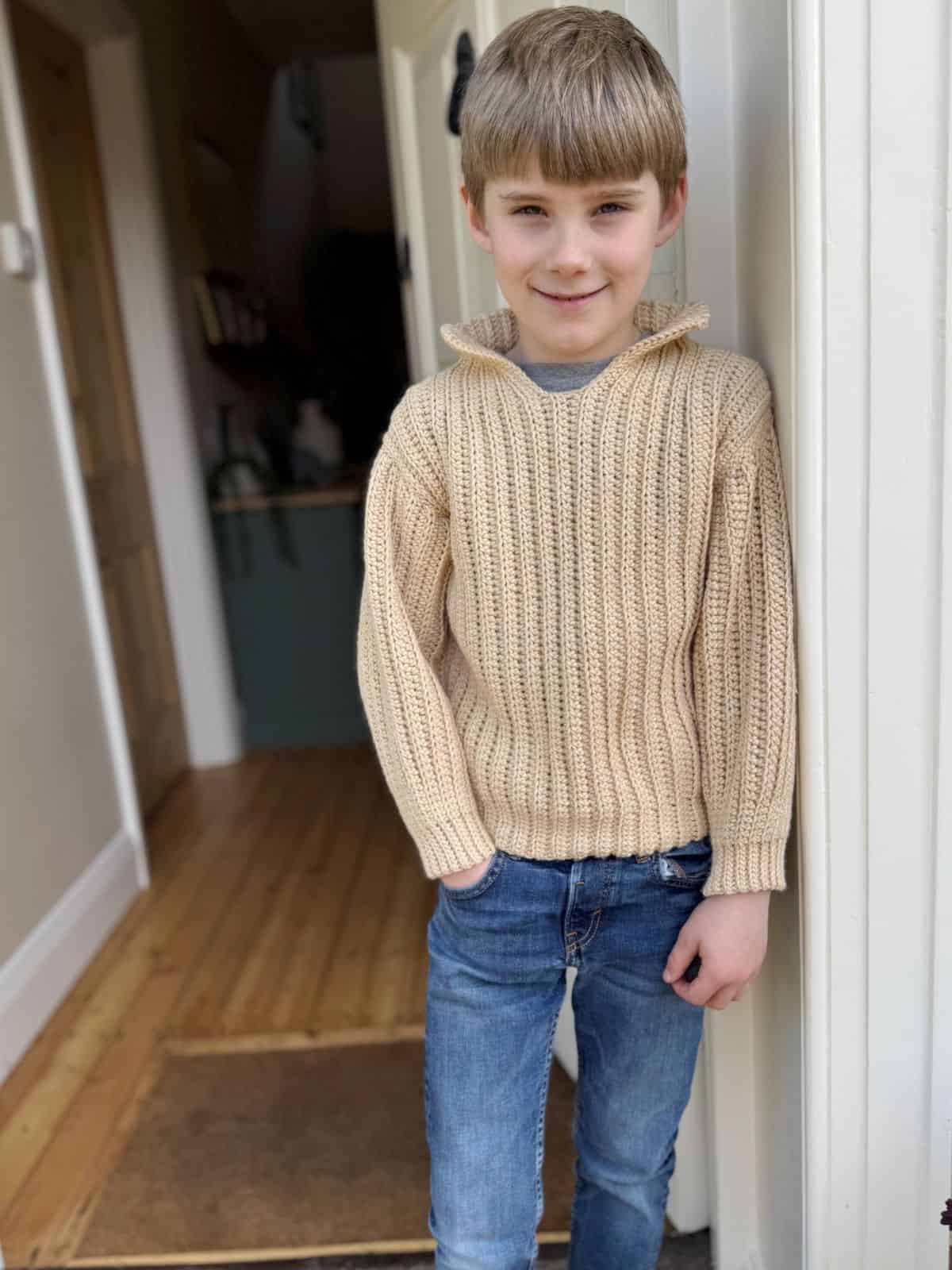 A young boy in a beige knit sweater and jeans stands in a doorway, smiling with hands in his pockets. His cozy sweater seems inspired by easy crochet patterns for beginners. The room has light wood flooring and partially visible furnishings.