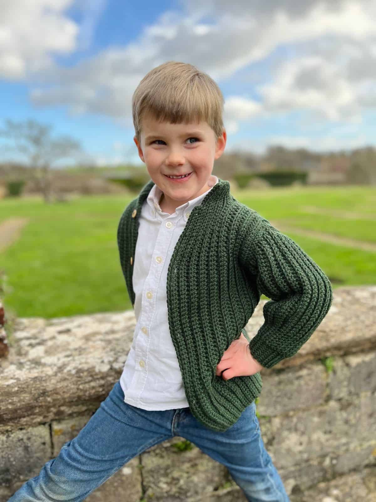 A young child with short hair poses outdoors, leaning against a stone wall with hands on hips. They wear a green knitted cardigan reminiscent of easy crochet sweater patterns for beginners, paired with a white shirt and blue jeans. A grassy landscape stretches out in the background.