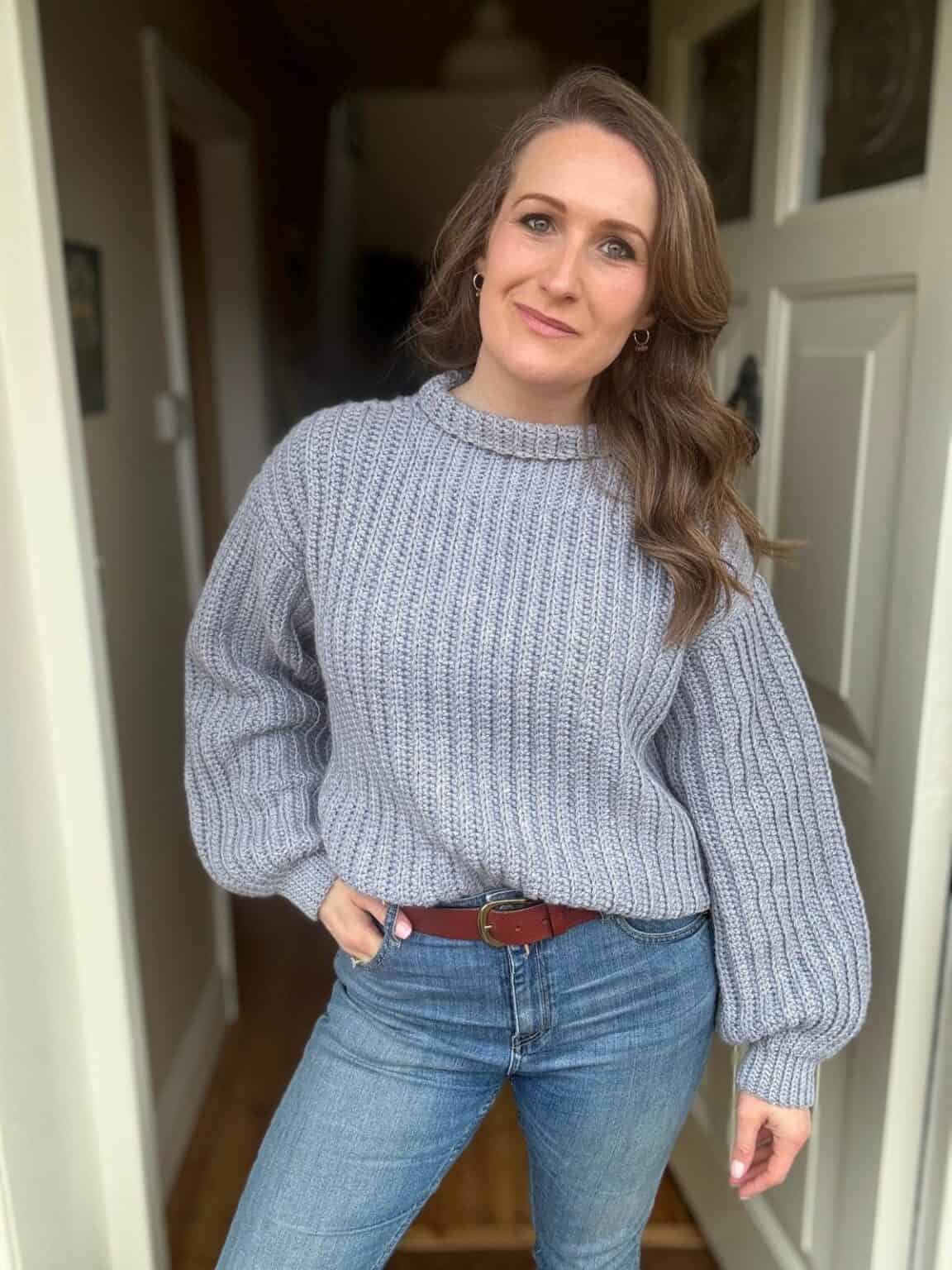 A person in blue jeans and an easy crochet sweater styled seamlessly for beginners stands with one hand on their hip, posing casually in a doorway.