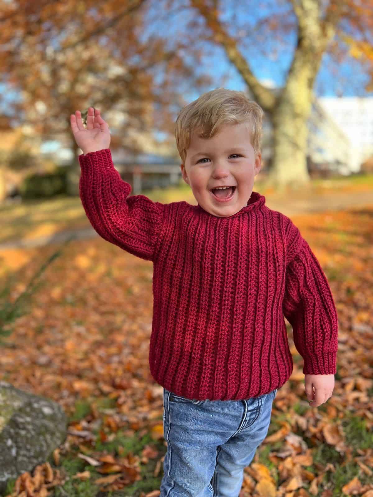 A young child in a cozy red sweater, reminiscent of easy crochet sweater patterns for beginners, smiles and waves while standing on a path adorned with fallen leaves. In the background, trees boast vibrant autumn foliage.