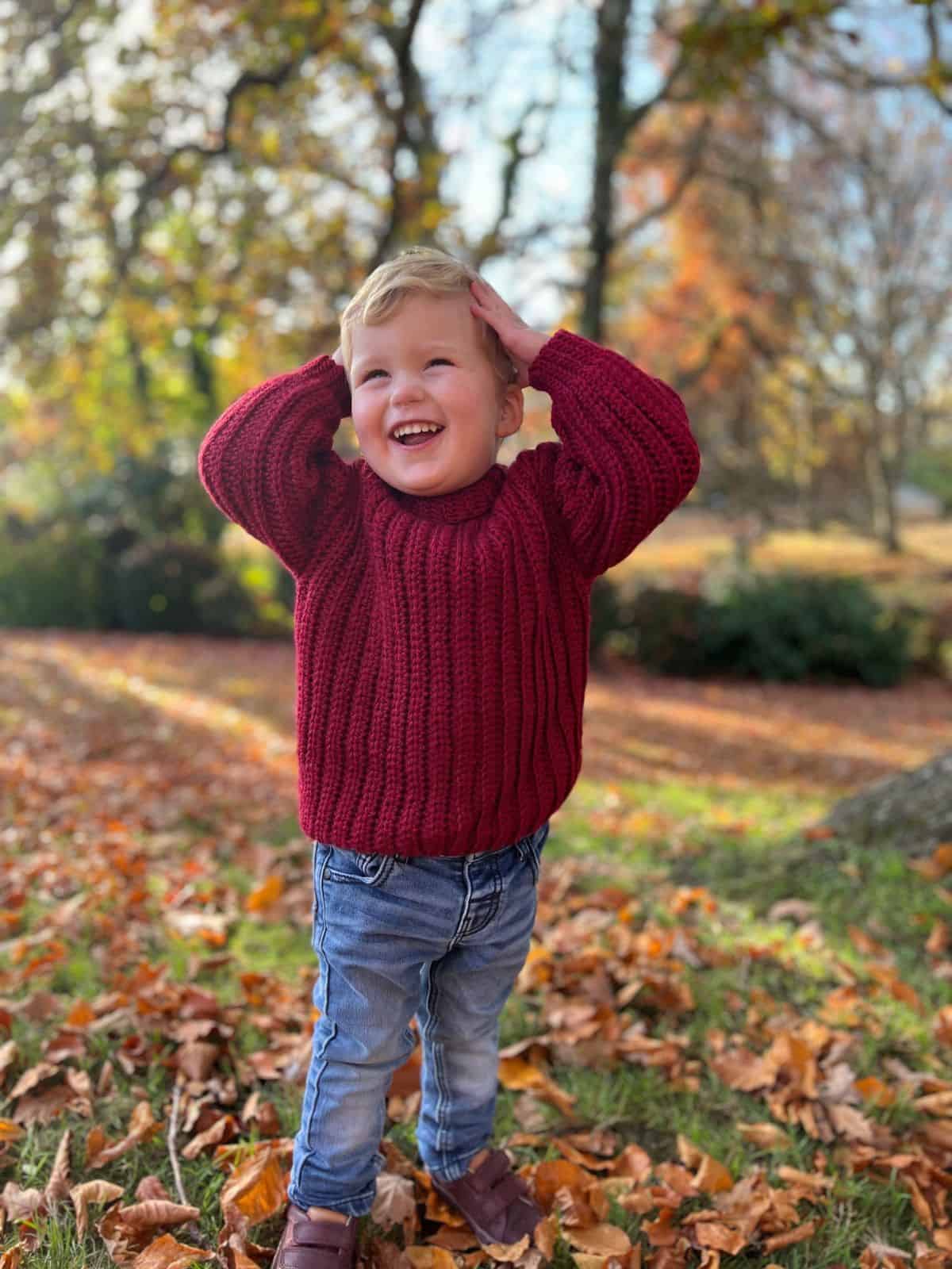 A child in a red sweater and jeans stands smiling with hands on head, surrounded by fallen autumn leaves. The cozy knit hints at easy crochet sweater patterns for beginners perfect for crafting that warm look.