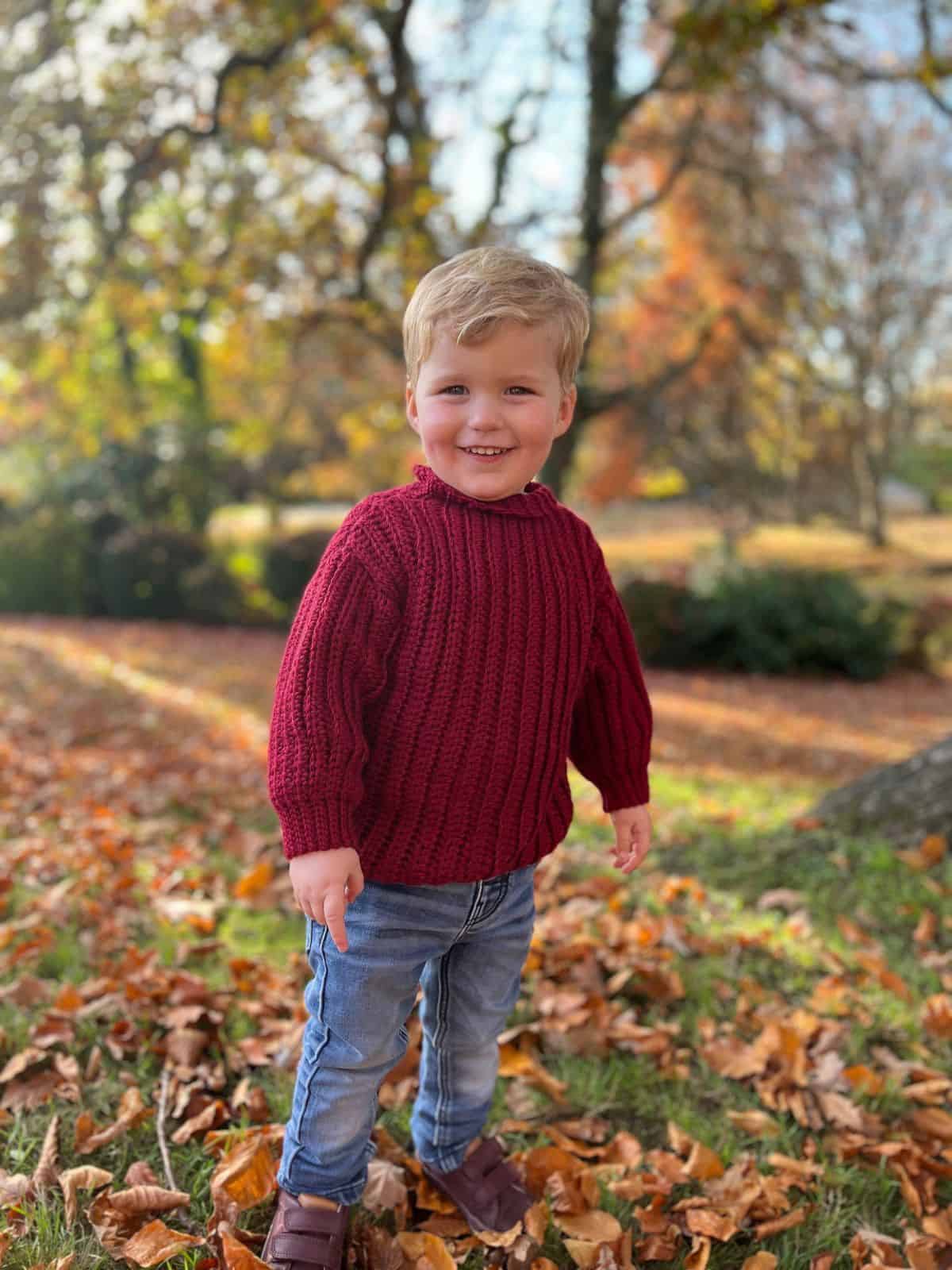 A smiling child stands on a grassy area covered with autumn leaves, wearing a vibrant red sweater reminiscent of easy crochet patterns for beginners, paired with jeans. Trees with fall foliage provide a stunning backdrop.