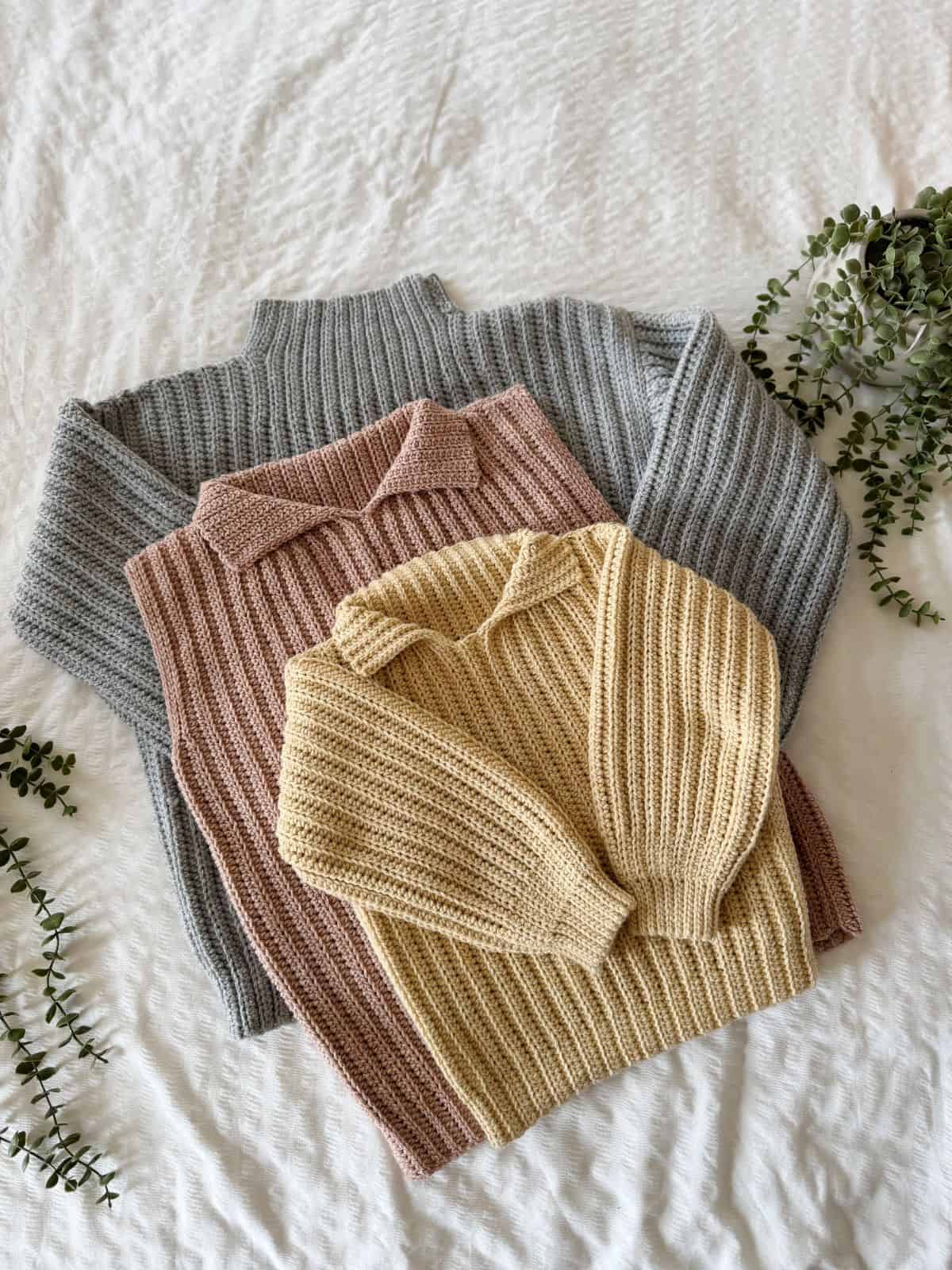 Three ribbed knit sweaters in gray, pink, and beige are neatly stacked on a soft textured surface, adorned with scattered green foliage—echoing the simplicity of easy crochet sweater patterns for beginners.