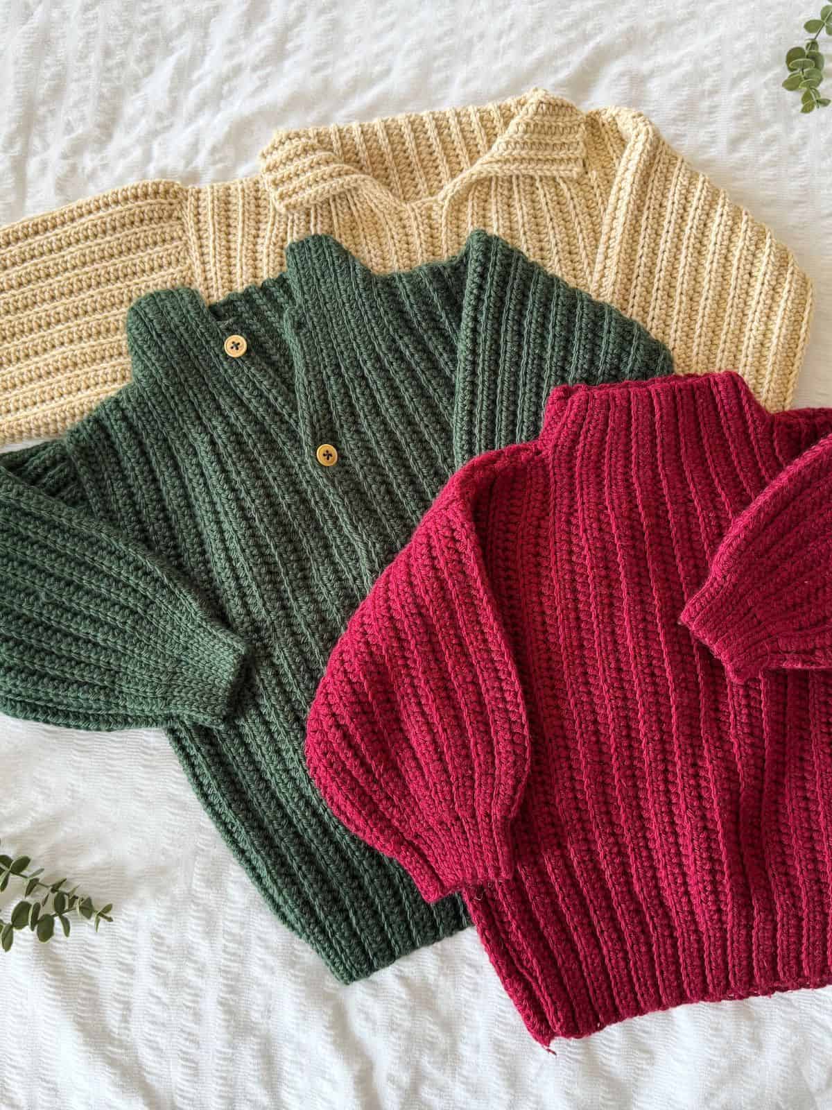 Three knitted sweaters in cream, forest green, and red are laid out on a textured white surface, surrounded by small green leaves. Perfect for those looking into easy crochet sweater patterns for beginners to add a cozy touch to any wardrobe.