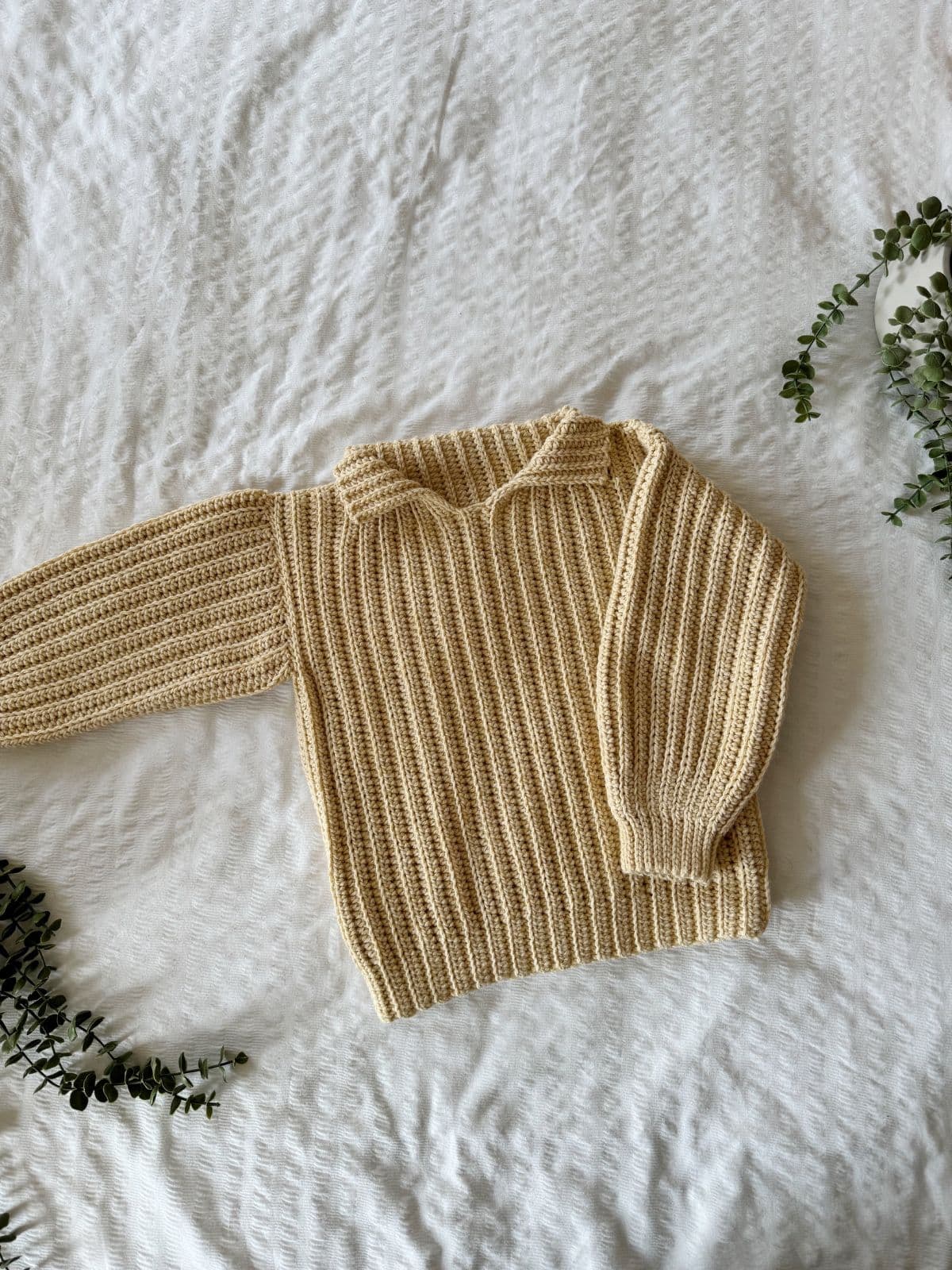 A cream-colored, ribbed knit sweater is laid on a textured white surface, surrounded by small green foliage accents, reminiscent of easy crochet sweater patterns for beginners.