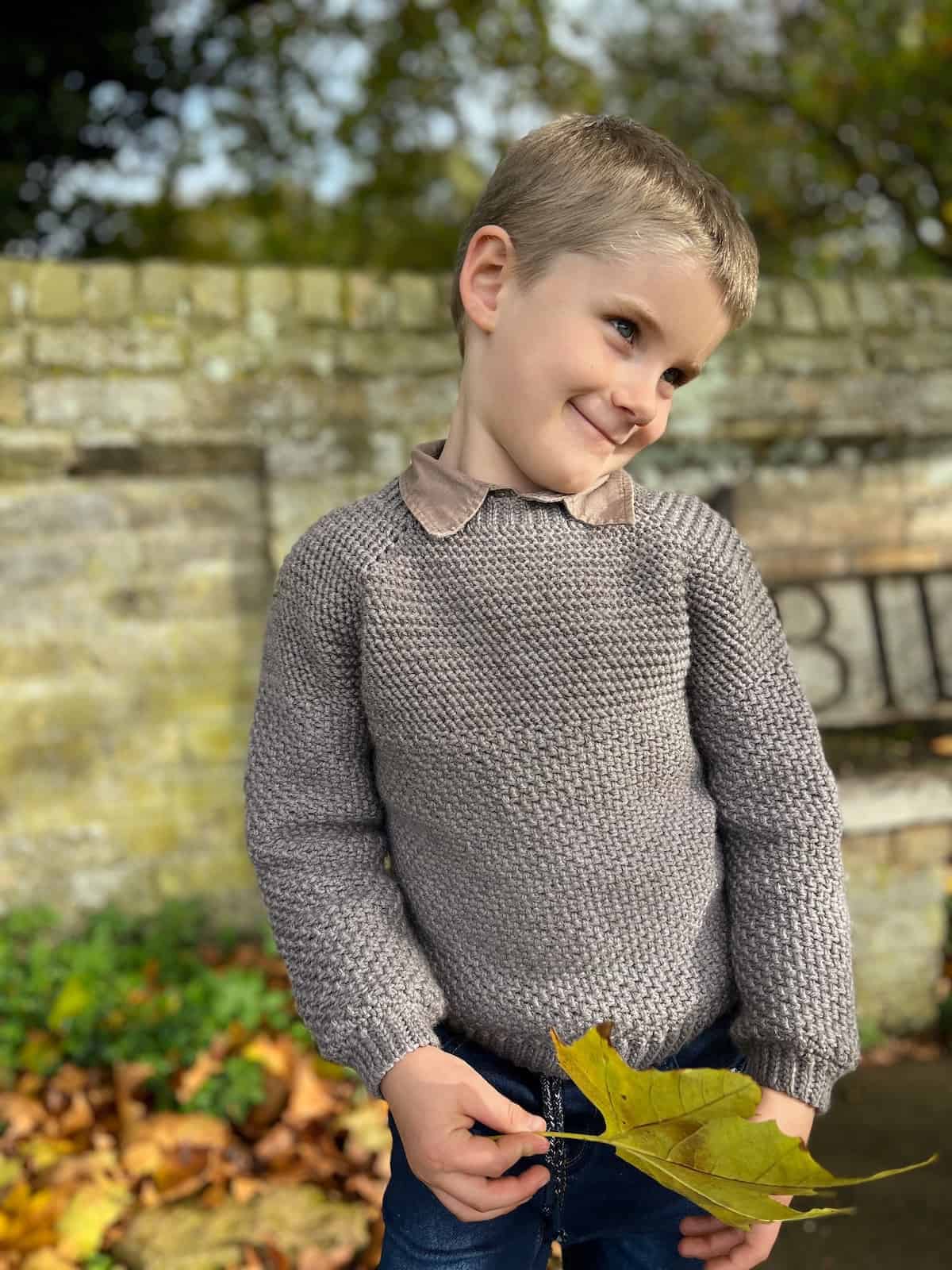 Boy wearing crochet sweater holding leaf and smiling.