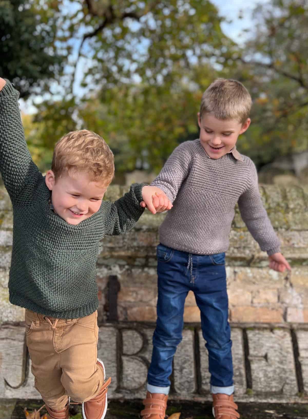 Two young boys, sporting cozy crochet sweaters and jeans, joyfully hold hands and balance on a ledge in a park. Trees and a stone wall are visible in the background.