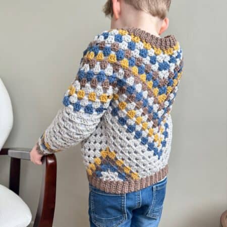 A young boy wearing a knitted sweater stands in front of a chair.