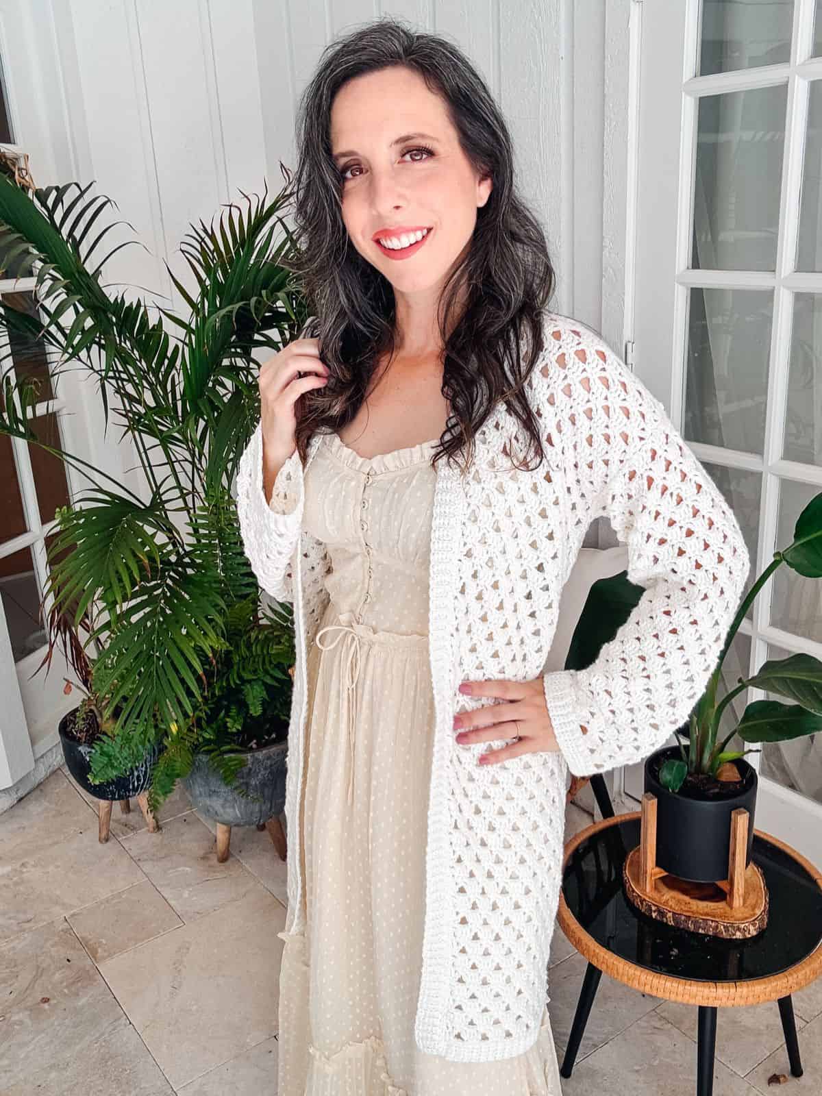 A woman with long dark hair stands indoors next to potted plants, wearing a beige dress and a white crocheted cardigan, smiling at the camera.