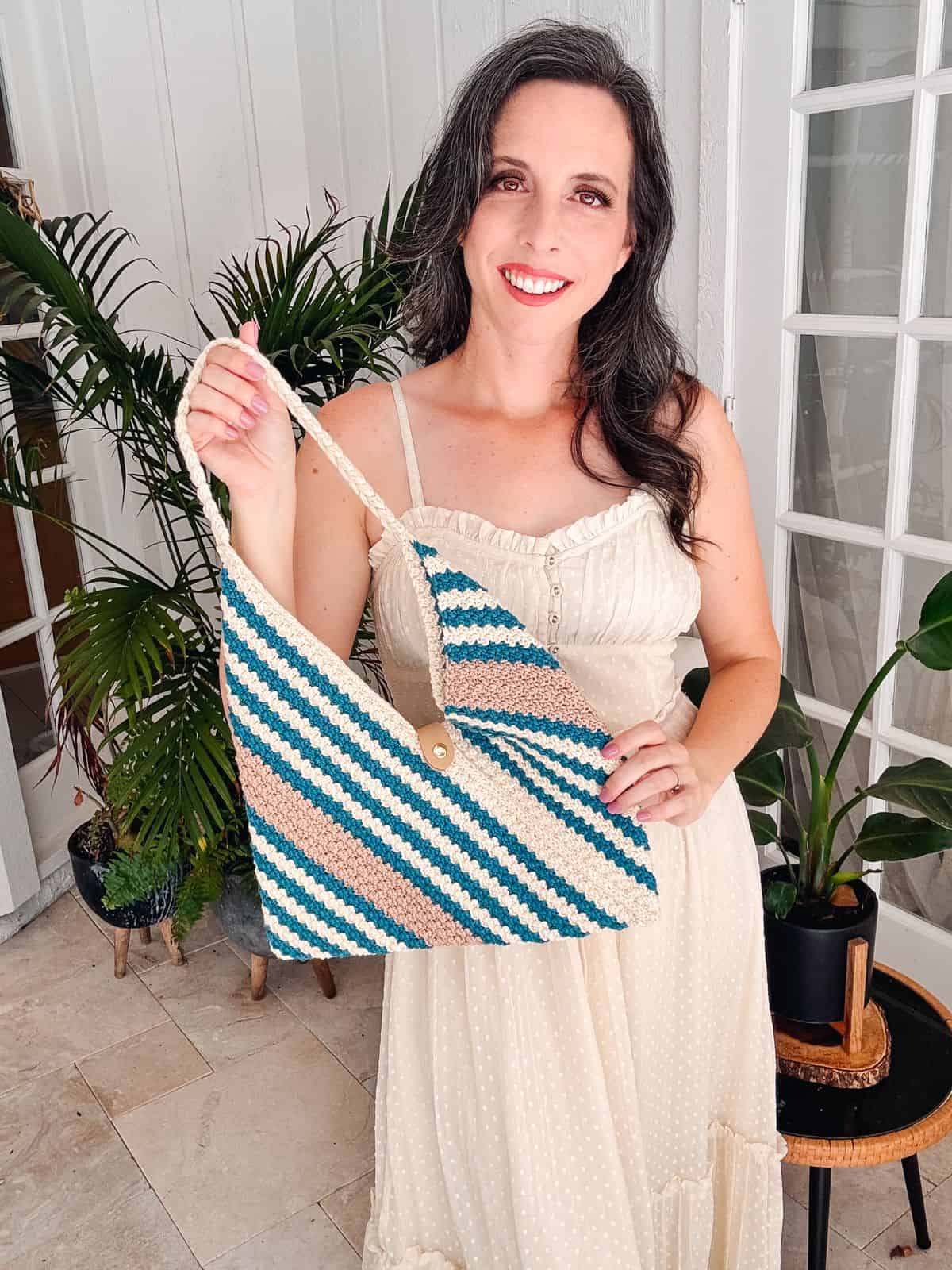 A woman in a cream dress smiles while holding a blue, white, and beige striped woven bag in a room with plants.