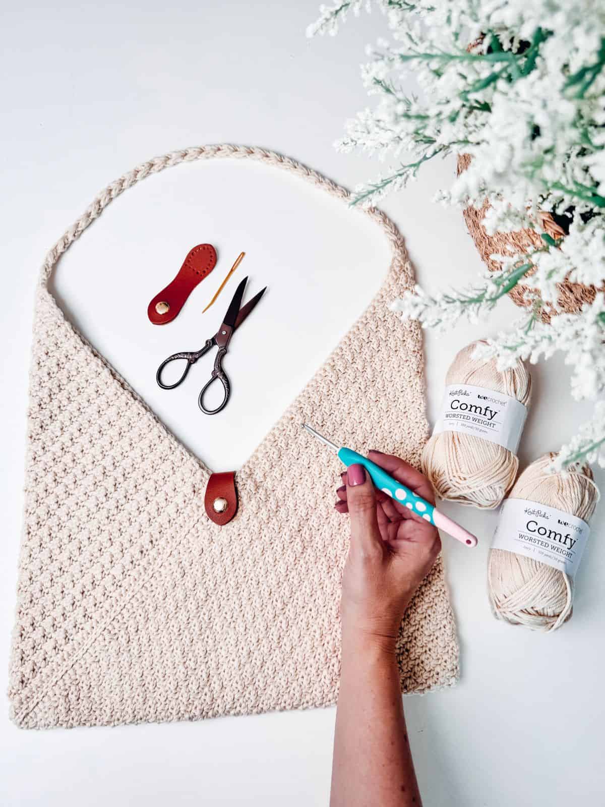 A person is crocheting with beige yarn next to a crocheted envelope-style bag, a pair of scissors, a crochet hook, and crochet tools on a white surface, with white flowers in the background.