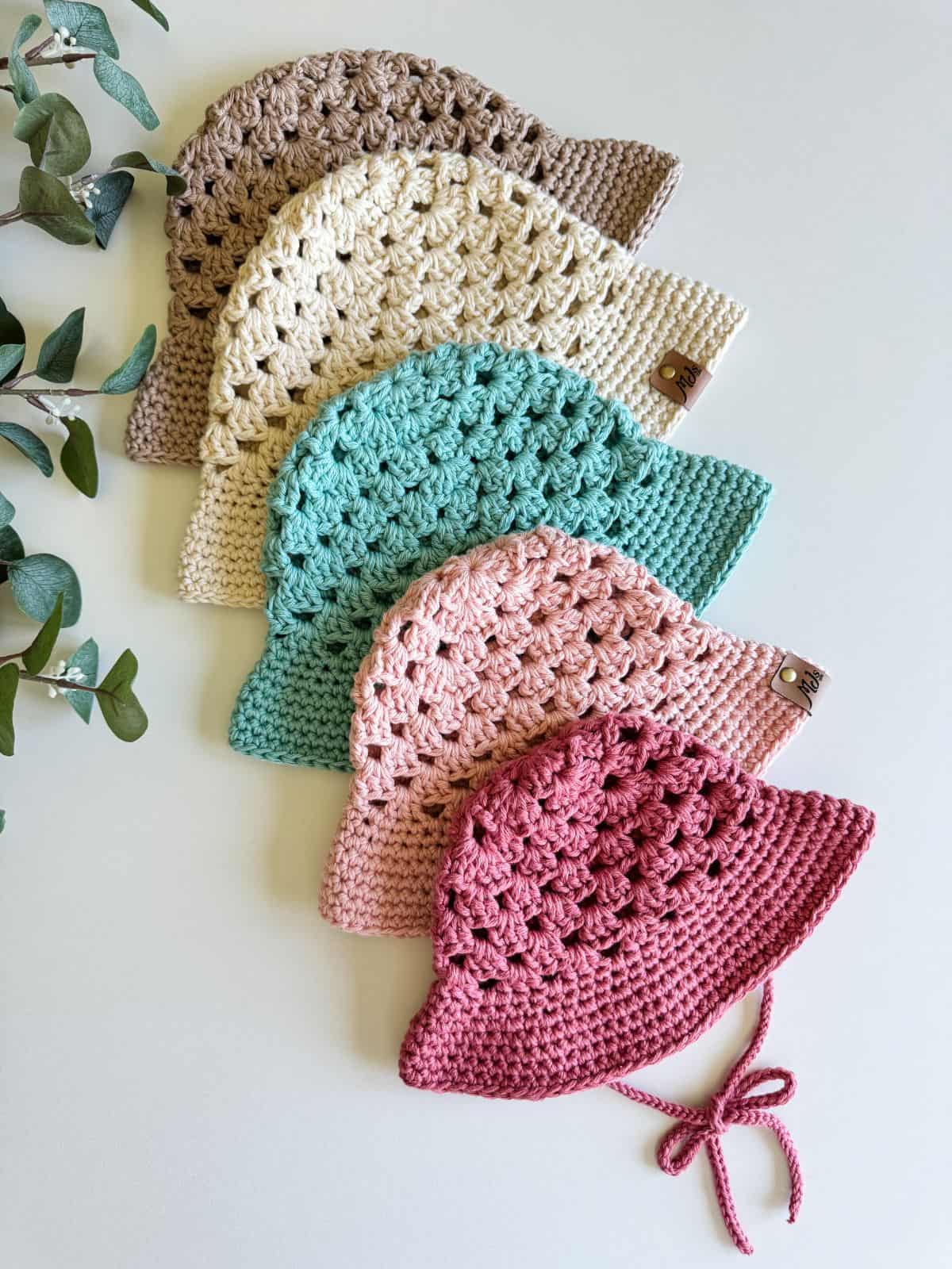 Five crocheted hats in varying colors (brown, cream, teal, light pink, and dark pink) are arranged neatly in a row. A leafy green plant is visible on the left side of the image.