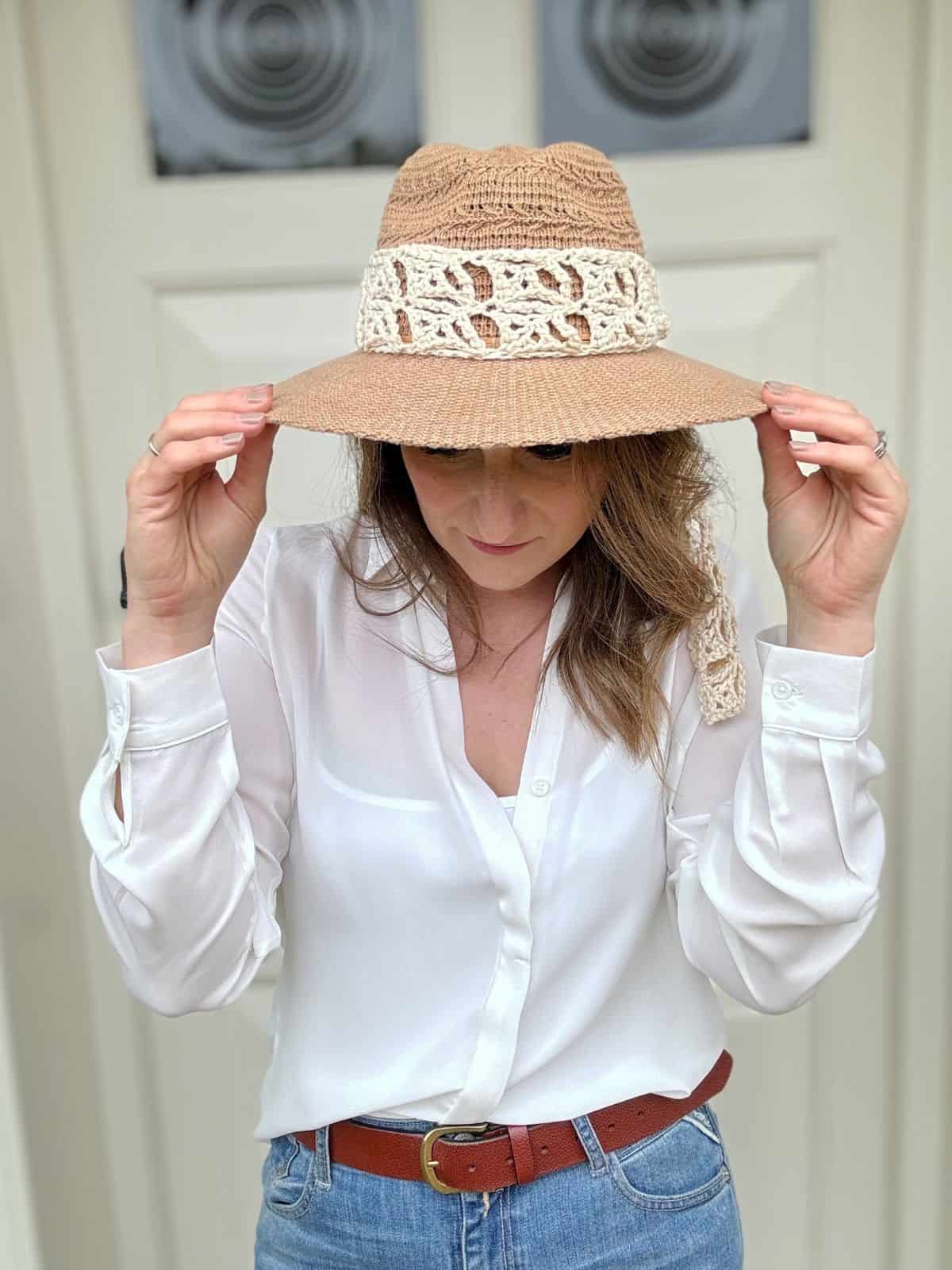 A person wearing a white blouse and jeans adjusts their beige hat with lace decorations in front of a white door.