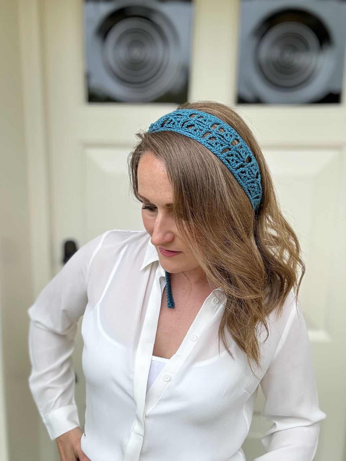 A woman with medium-length, wavy hair wears a blue lace headband and a white blouse. She is looking down, standing in front of a door with circular designs on the glass panels.
