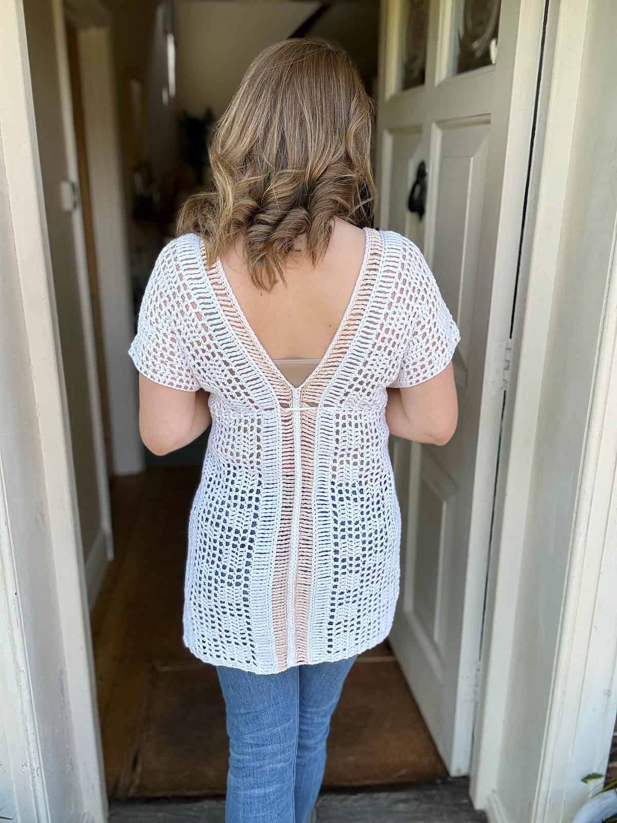 Rear view of a person with shoulder-length hair wearing a white crochet bikini top pattern over a beige undershirt and blue jeans, standing inside a doorway.