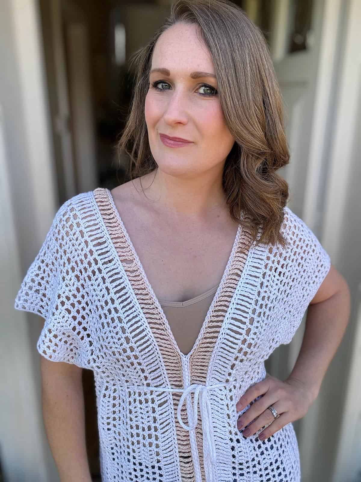 A person with light brown hair wearing a white crochet top stands indoors with a neutral expression, perhaps contemplating the next crochet bikini top pattern to craft.