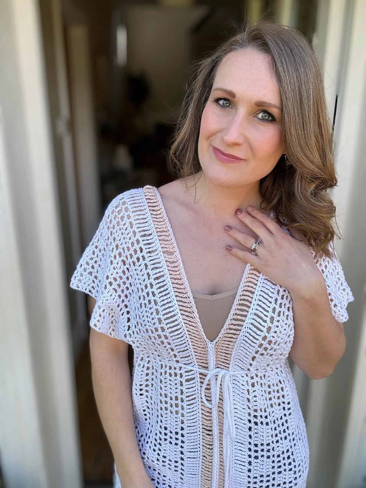 A woman stands in a doorway, wearing a white crocheted top that looks like it was inspired by a crochet bikini top pattern. She has wavy, shoulder-length hair and rests her right hand on her chest, displaying a ring.