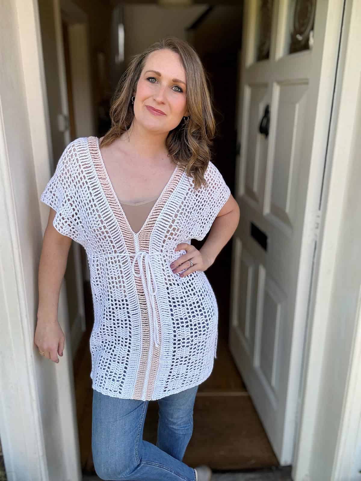 Woman standing in an open doorway, wearing a white crochet top over a tan garment, and blue jeans, with one hand resting on the door frame. The intricate design looks like it could be from a crochet bikini top pattern, adding to her casual yet chic appearance.