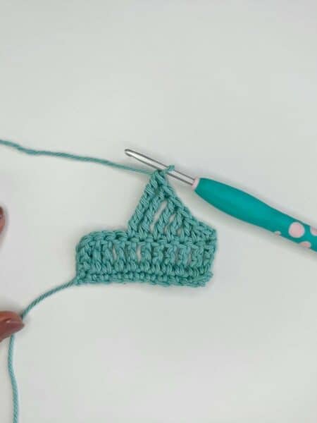 A hand with painted nails holds the edge of a light blue crochet piece in progress, featuring long crochet stitches. A crochet hook with a blue and pink handle is attached to the yarn beside the piece.