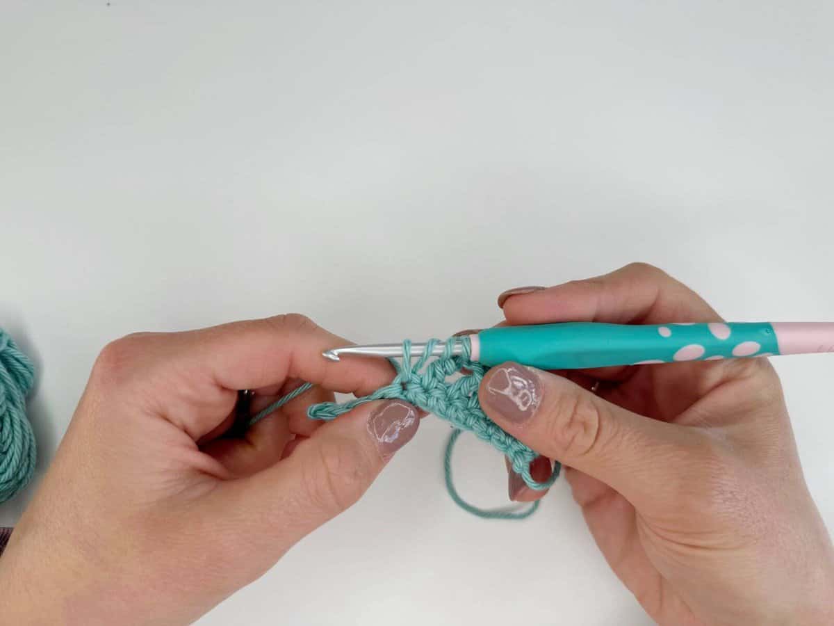 Hands are shown holding a knitting project in progress with a turquoise crochet hook. The long crochet stitches add a touch of elegance, perfectly complementing the pale pink polish with white floral designs on the fingernails.