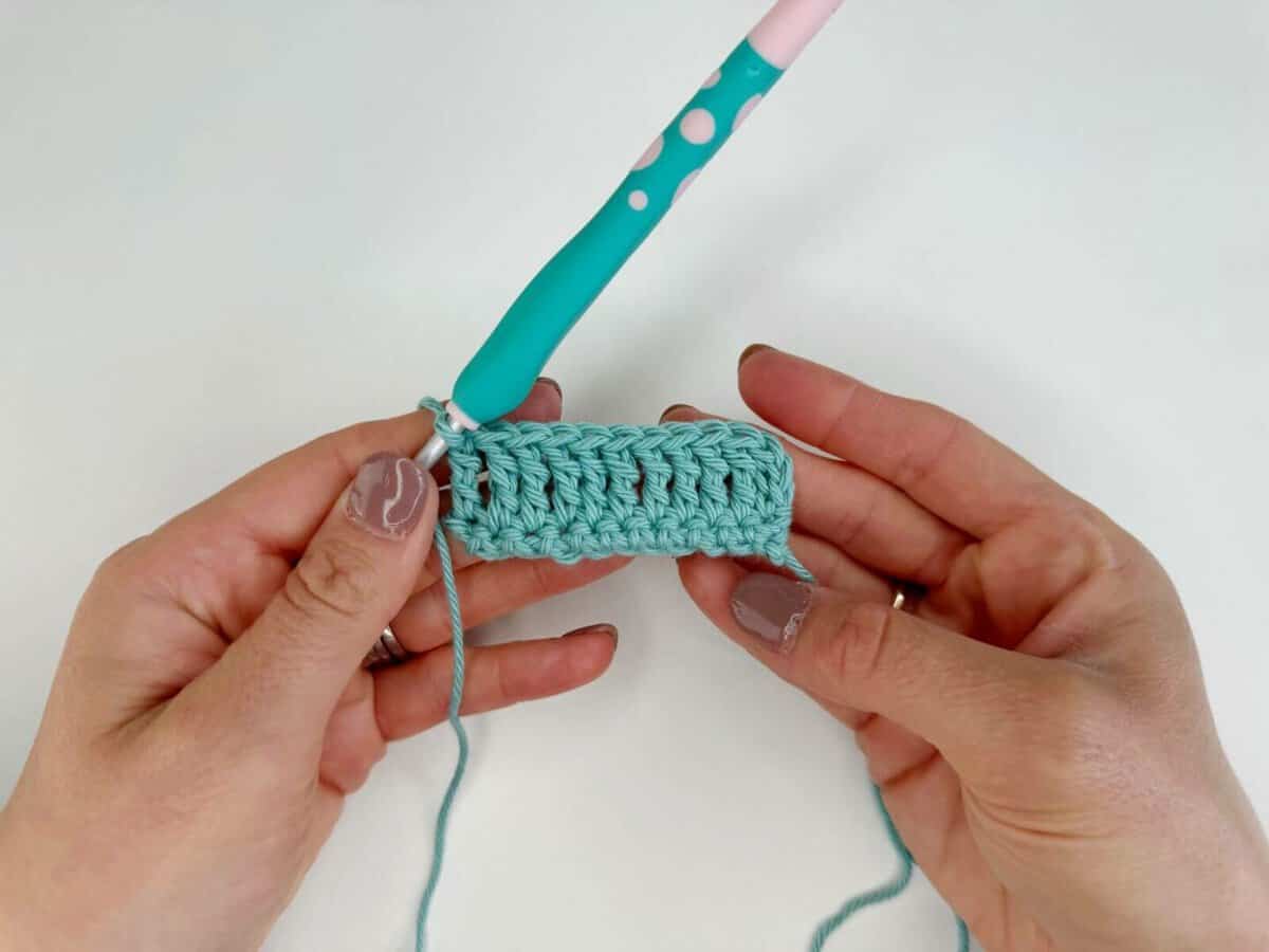 Hands holding a crocheted piece with long crochet stitches and a turquoise crochet hook with a pink handle.