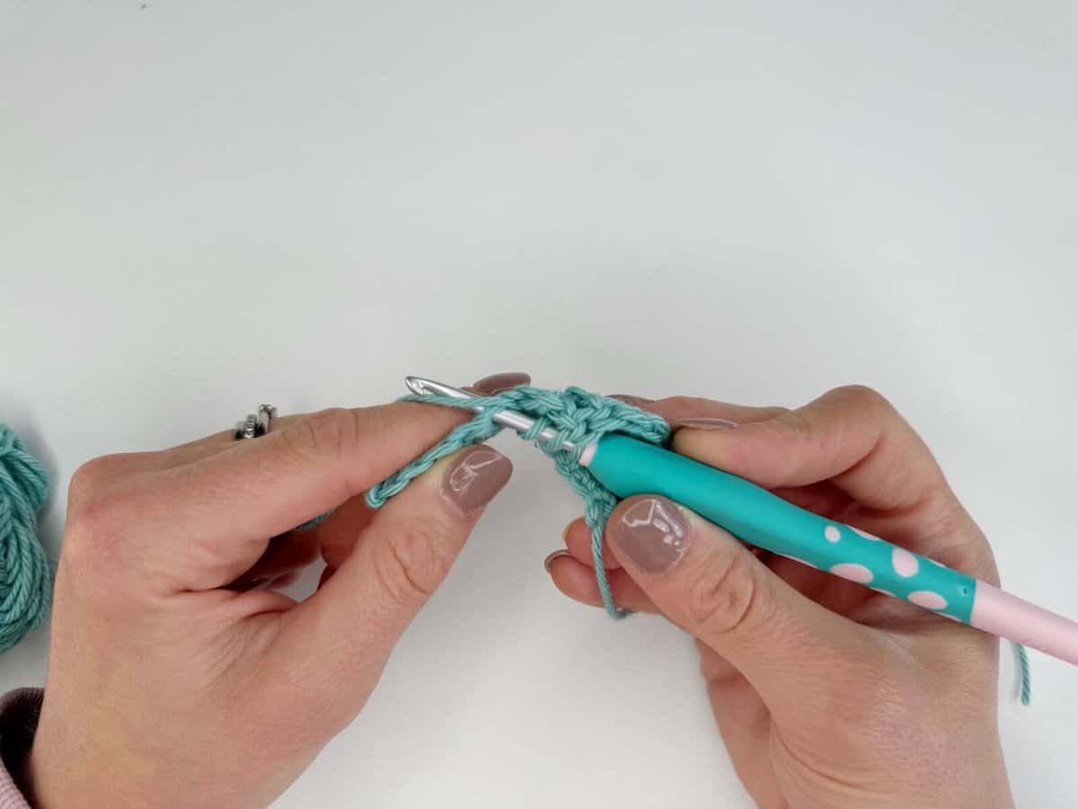 Hands crocheting with a teal and pink crochet hook, working long crochet stitches with light blue yarn against a white background.