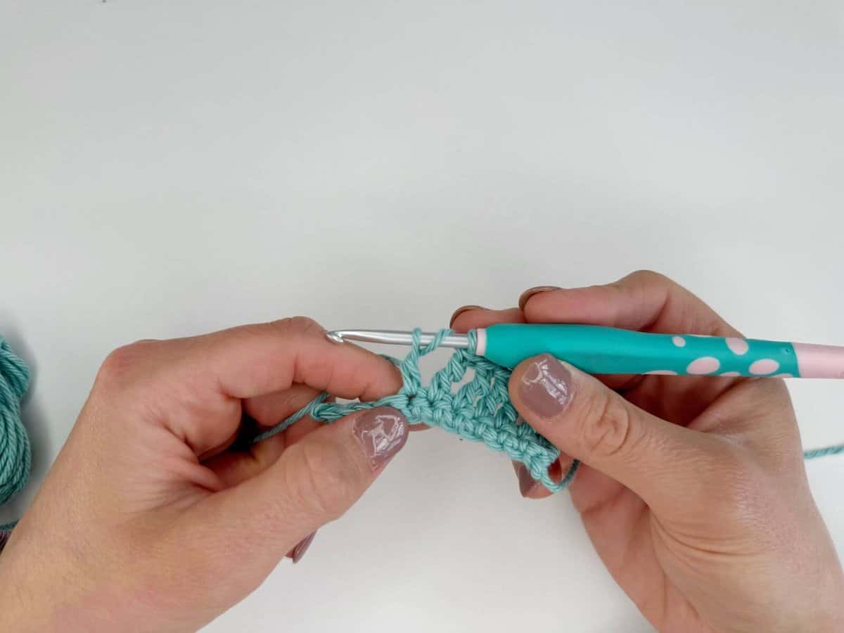 Two hands are crocheting with a teal yarn and a green crochet hook, creating long crochet stitches on a white background.