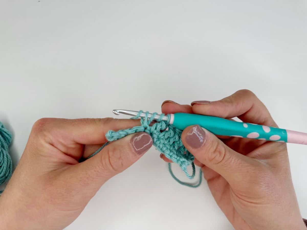 Two hands creating long crochet stitches with light teal yarn and a multicolored crochet hook against a plain white background.