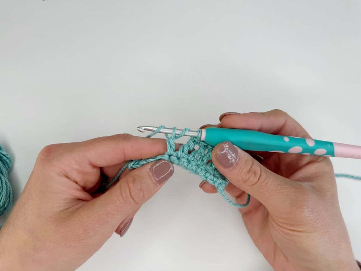 Two hands are expertly crocheting with a teal yarn and a crochet hook that has a pink and white polka dot handle, creating long crochet stitches with precision.