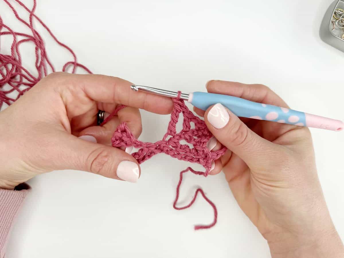 A person is crocheting with a pink yarn and a crochet hook with a pink and blue handle. The hands hold a partially completed crochet piece with long crochet stitches against a white background.