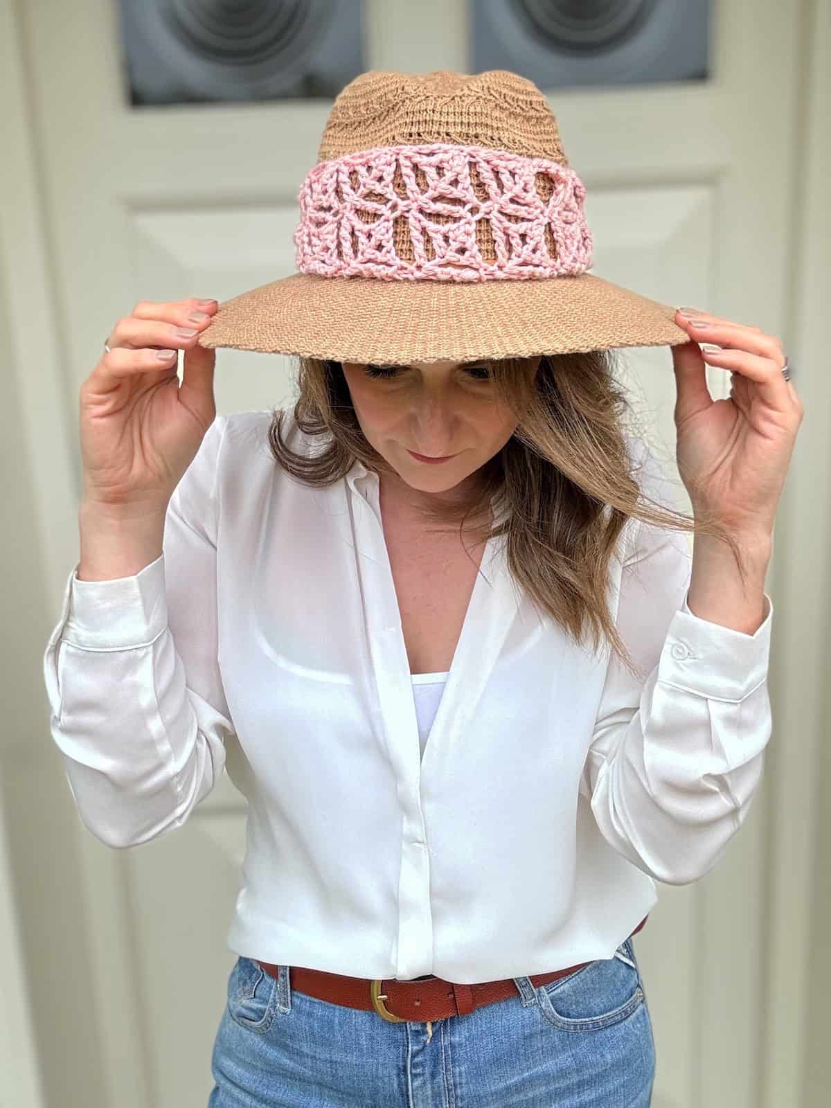 A person wearing a white shirt, blue jeans, and a wide-brimmed hat with a pink lace crochet hat band adjusts the brim while looking down.