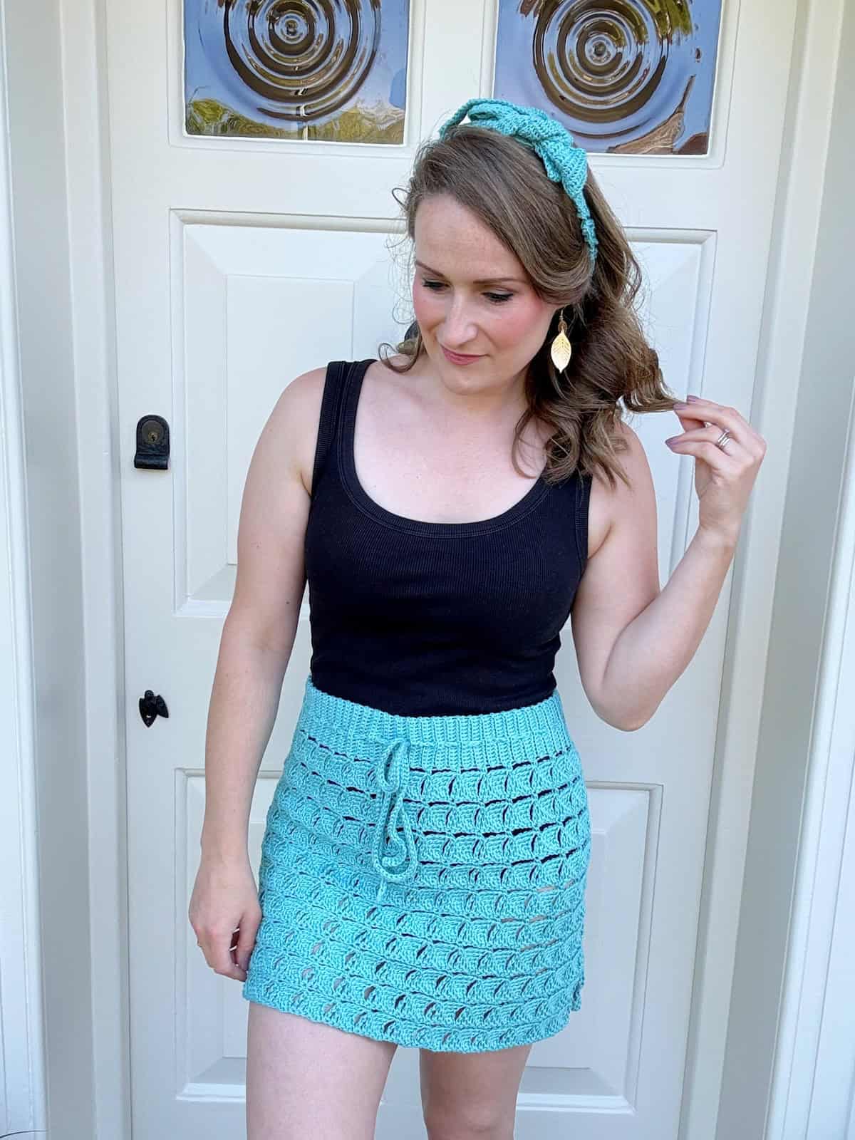 A woman stands in front of a white door wearing a black tank top and a turquoise crocheted skirt with a matching headband, looking down and to the side, touching her hair with her right hand. She seems to be admiring the intricate crochet skirt pattern she crafted. 