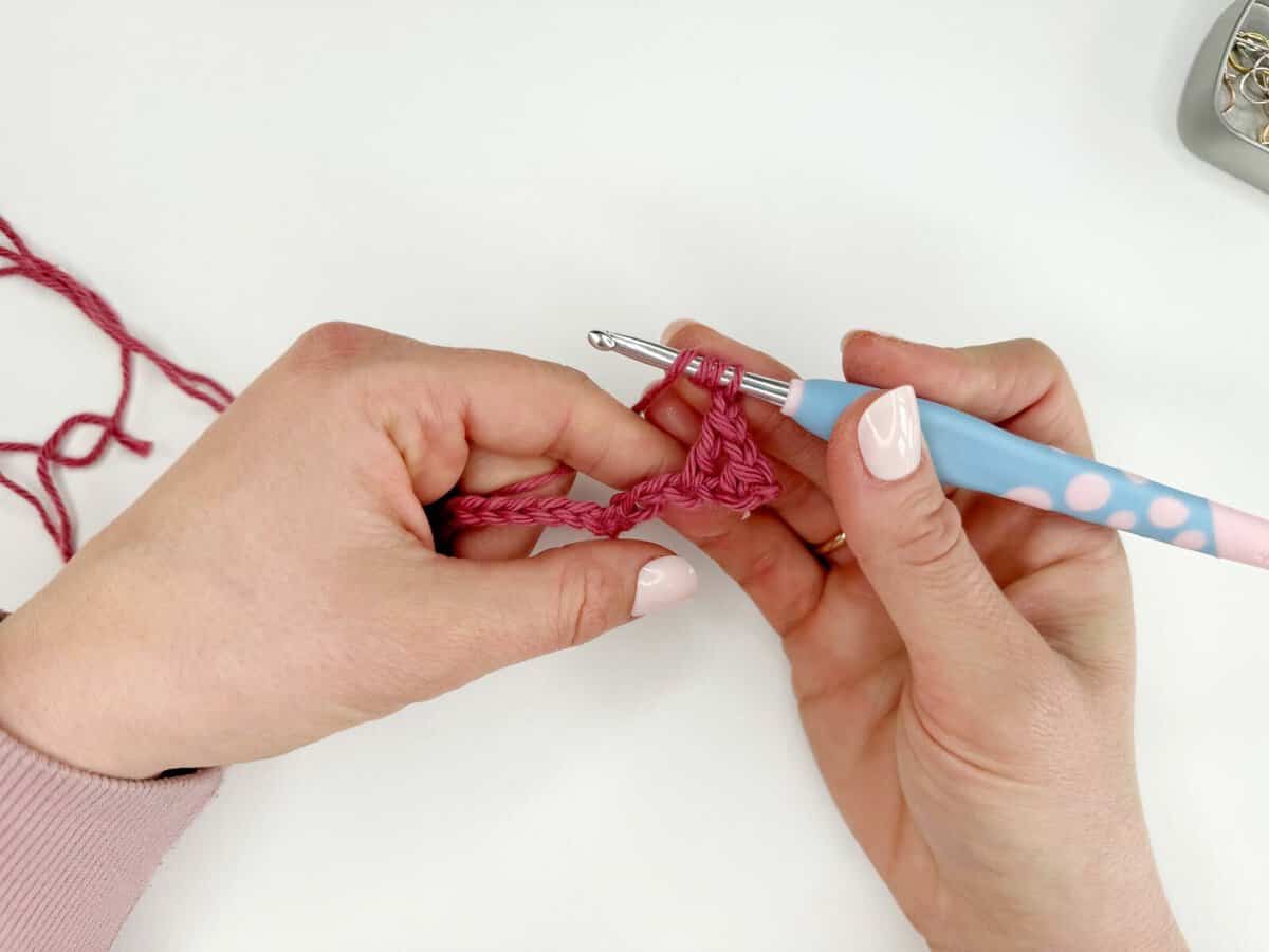 Close-up of hands crocheting long stitches with pink yarn and a blue crochet hook against a white background.