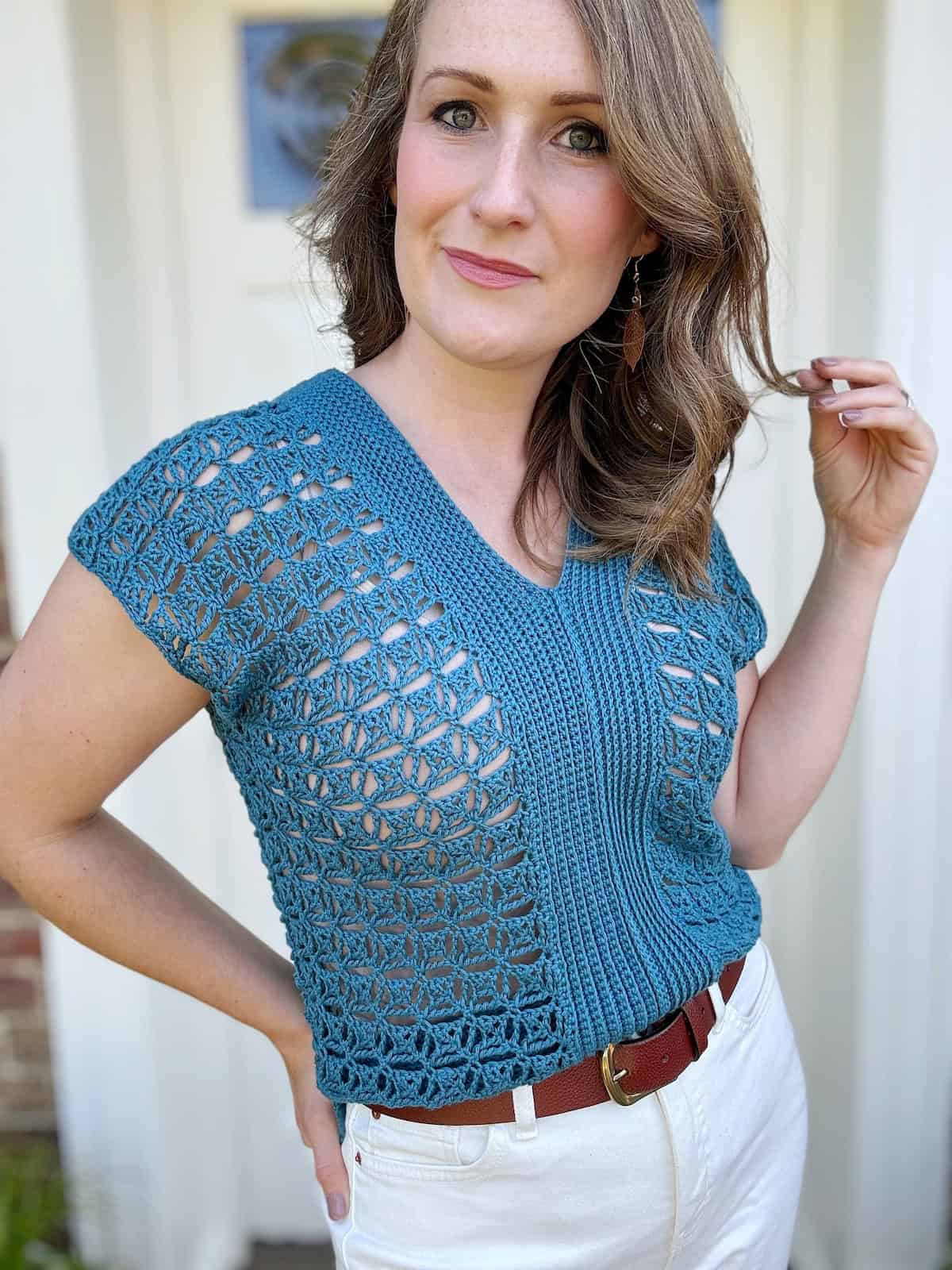 A woman stands in front of a white door, wearing a blue crocheted top with geometric patterns and white pants, holding a strand of her hair with a slight smile.