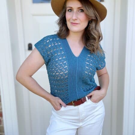 Person standing in front of a door, wearing a blue crocheted top, white pants, brown belt, and a straw hat.